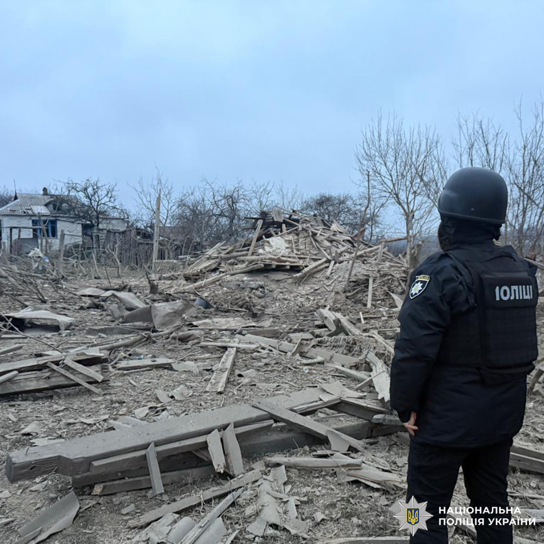 Поліцейський стоїть серед руїн після масштабного руйнування. Навколо уламки дерев’яних конструкцій, повалені дерева та пошкоджена будівля на тлі.