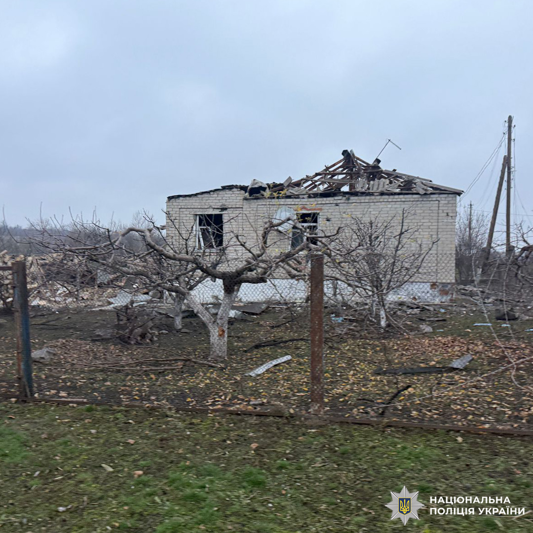 Одноповерховий цегляний будинок із частково зруйнованим дахом, ймовірно пошкоджений унаслідок стихійного лиха або бойових дій. На подвір’ї уламки, голі дерева та огорожа.