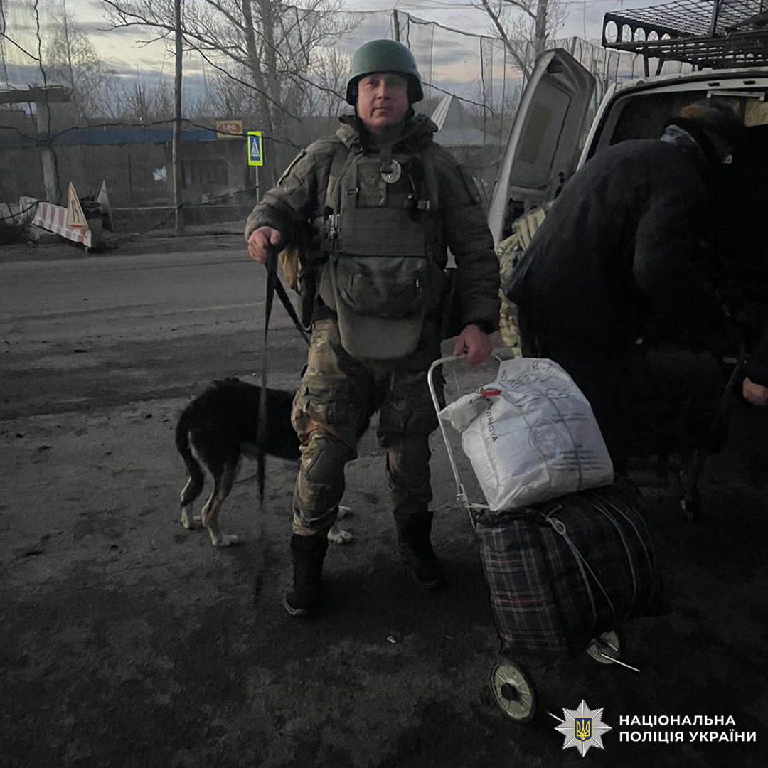 Поліцейський у камуфляжі та бронежилеті стоїть на дорозі поруч із транспортом, тримаючи палицю та візок із речами.