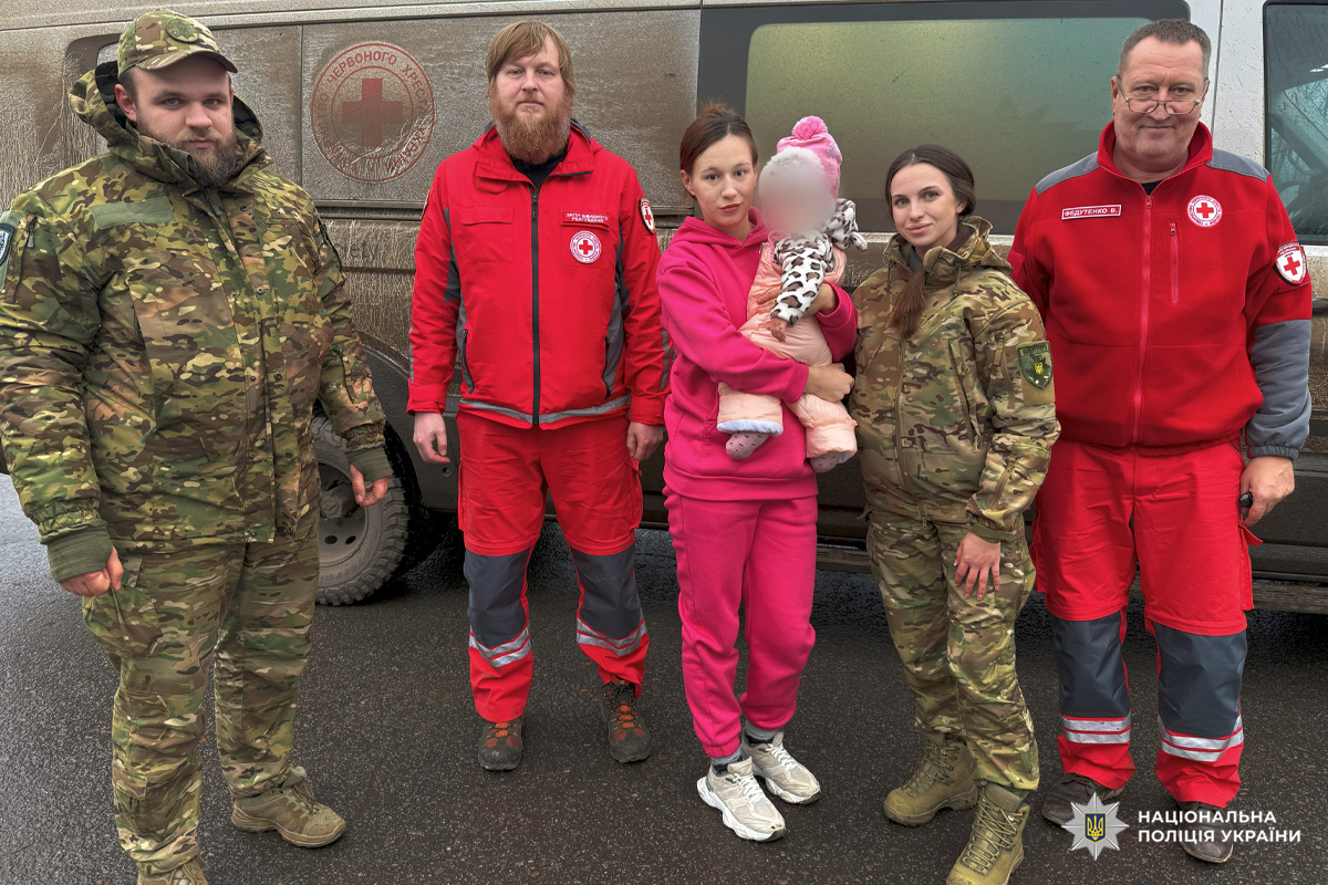 Шестеро людей біля автомобіля: двоє у формі медичних служб з емблемами Червоного Хреста, двоє  поліцейських у камуфляжі. Жінуа тримає малу дитину в рожевому одязі.