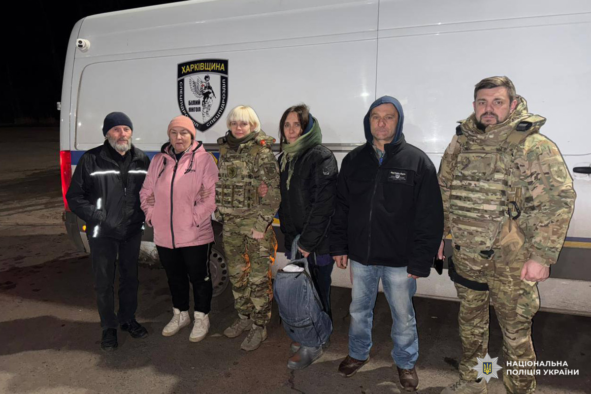 Шестеро людей біля білого фургона: двоє у поліцейських у камуфляжі, четверо у цивільному одязі. Один тримає сумку.