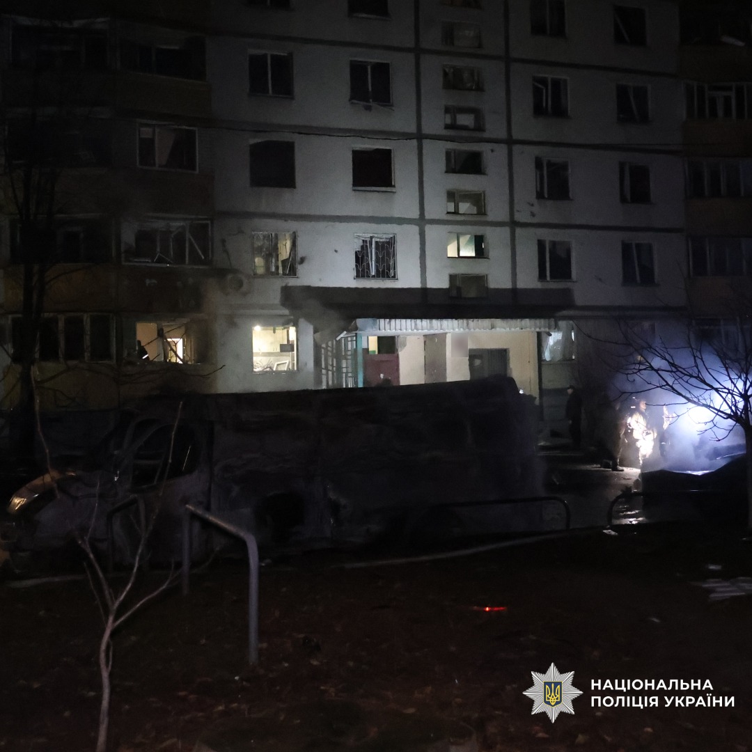 Нічна сцена біля пошкодженого житлового будинку. Вигорілий автомобіль, уламки, розбиті вікна. Освітлення від ліхтаря та внутрішнього світла.