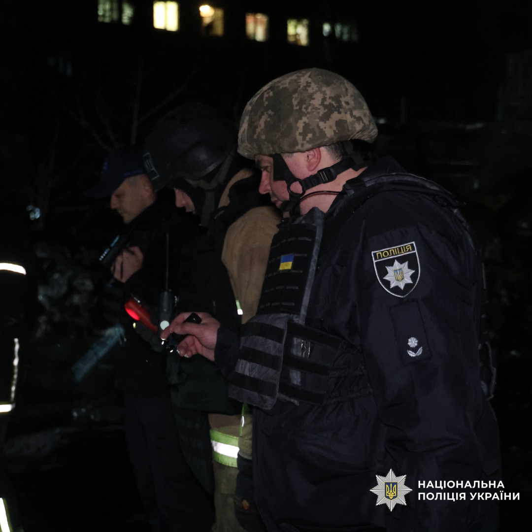 Правоохоронці та рятувальники працюють у темну пору доби. Один із поліцейських у бронежилеті з українським шевроном оглядає місце події з ліхтарем. На тлі будівля з освітленими вікнами.