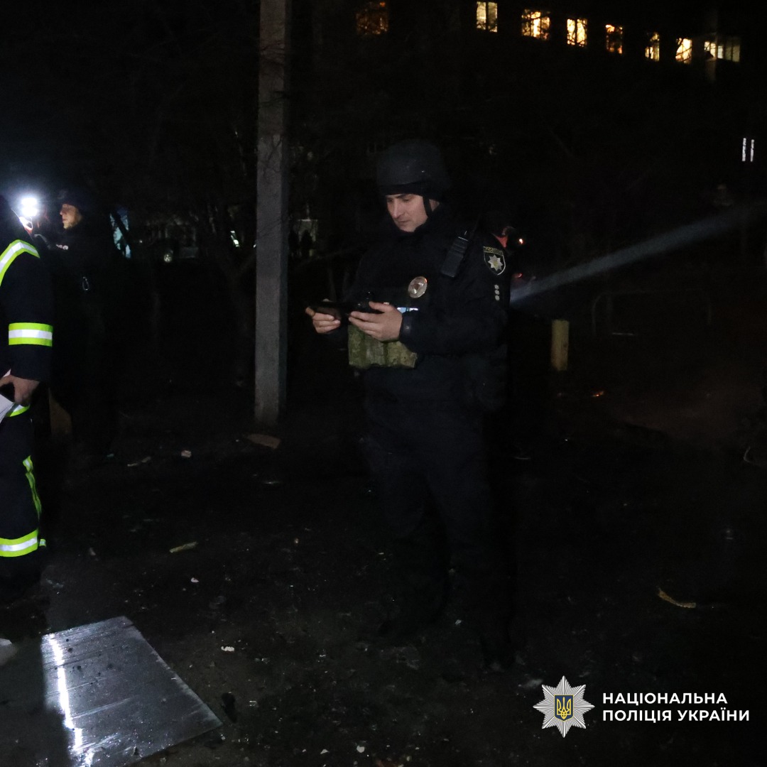 Нічна робота правоохоронців та рятувальників. Поліцейський у бронежилеті та шоломі тримає документ або пристрій. Мокра земля, уламки, освітлені вікна будівлі.