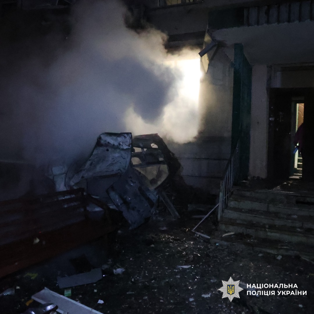 Наслідки вибуху біля входу в будівлю. Дим, уламки, пошкоджені конструкції. Відчинені двері, світло з вікна.