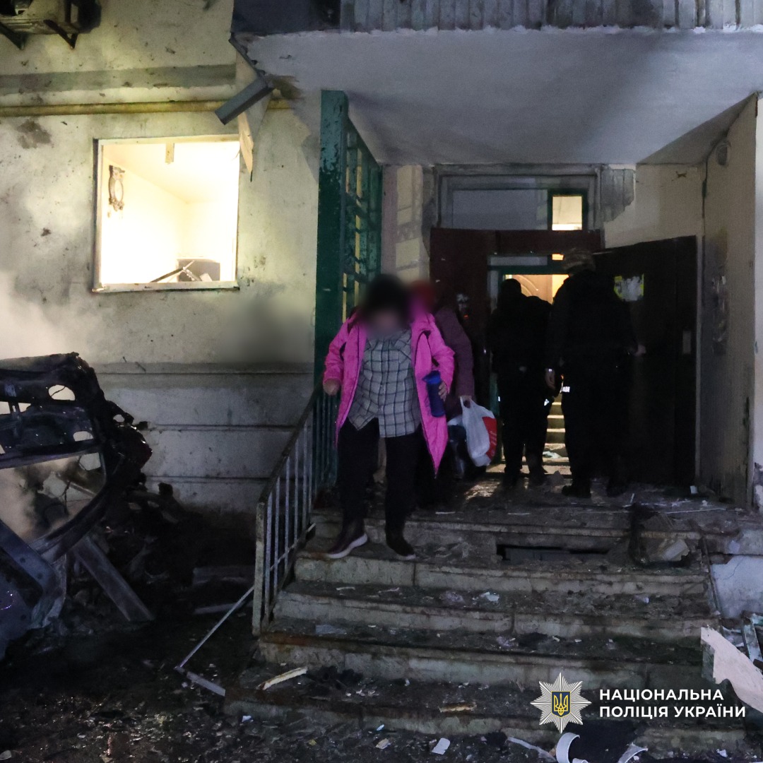 Пошкоджений вхід до будівлі, уламки, знищений транспорт. Люди виходять із приміщення, одна особа в рожевому пальті.