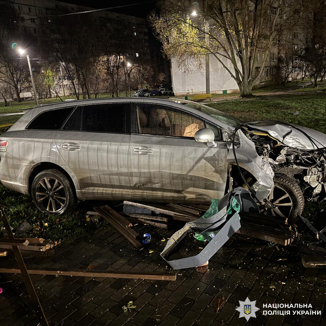 Передня частина авто сильно пошкоджена: капот зім’ятий, бампер і двигун деформовані. Навколо уламки автомобіля та конструкції. Подія сталася вночі, ймовірно, в парку або житловому районі.