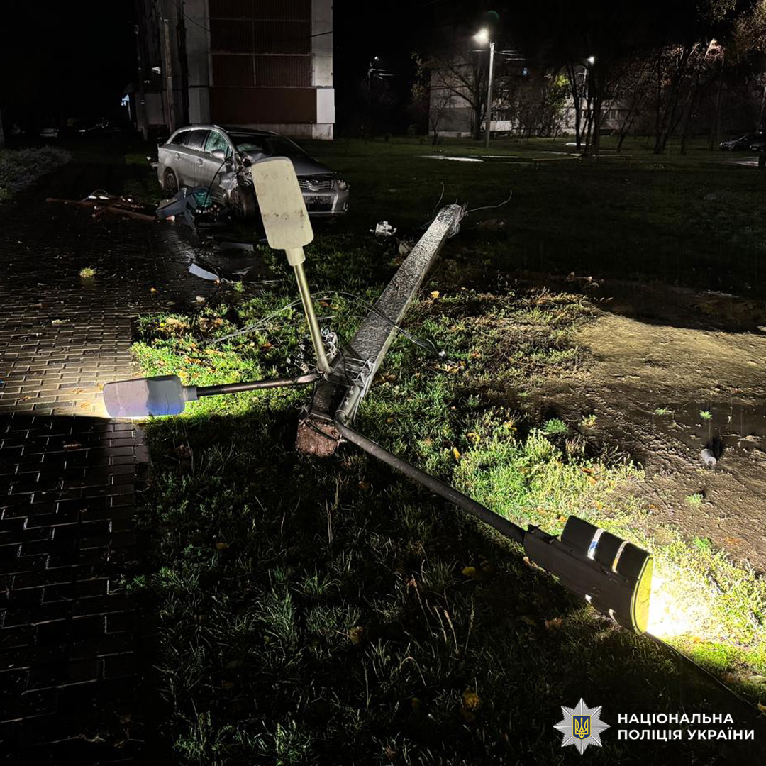 Автомобіль зніс опору вуличного освітлення, яка впала на газон біля пішохідної зони. Стовп зламаний біля основи, навколо уламки авто та конструкції. На задньому плані пошкоджений транспортний засіб.