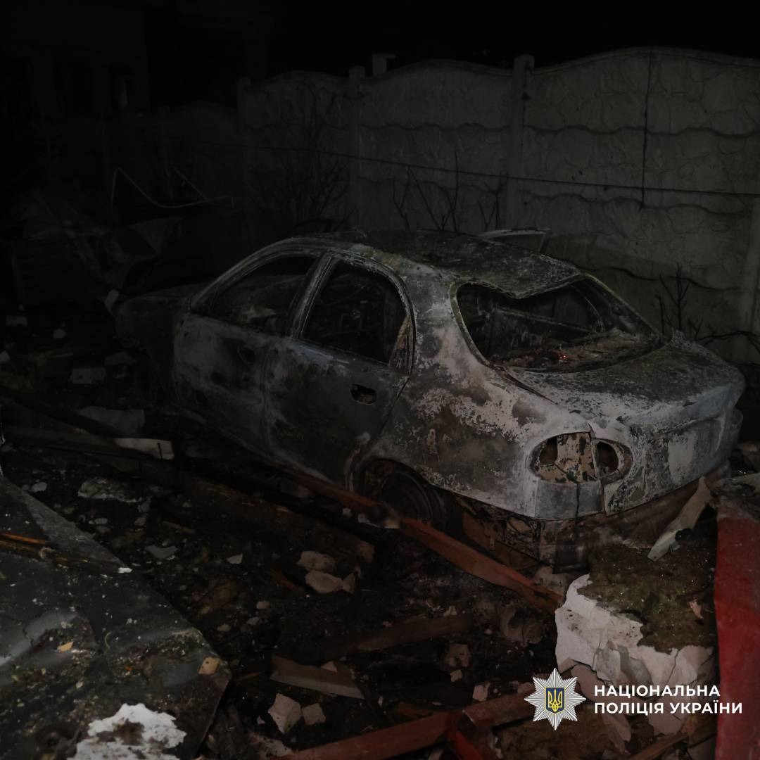 Обгорілий і зруйнований автомобіль у темному середовищі, навколо уламки дерева та пошкоджена бетонна стіна.