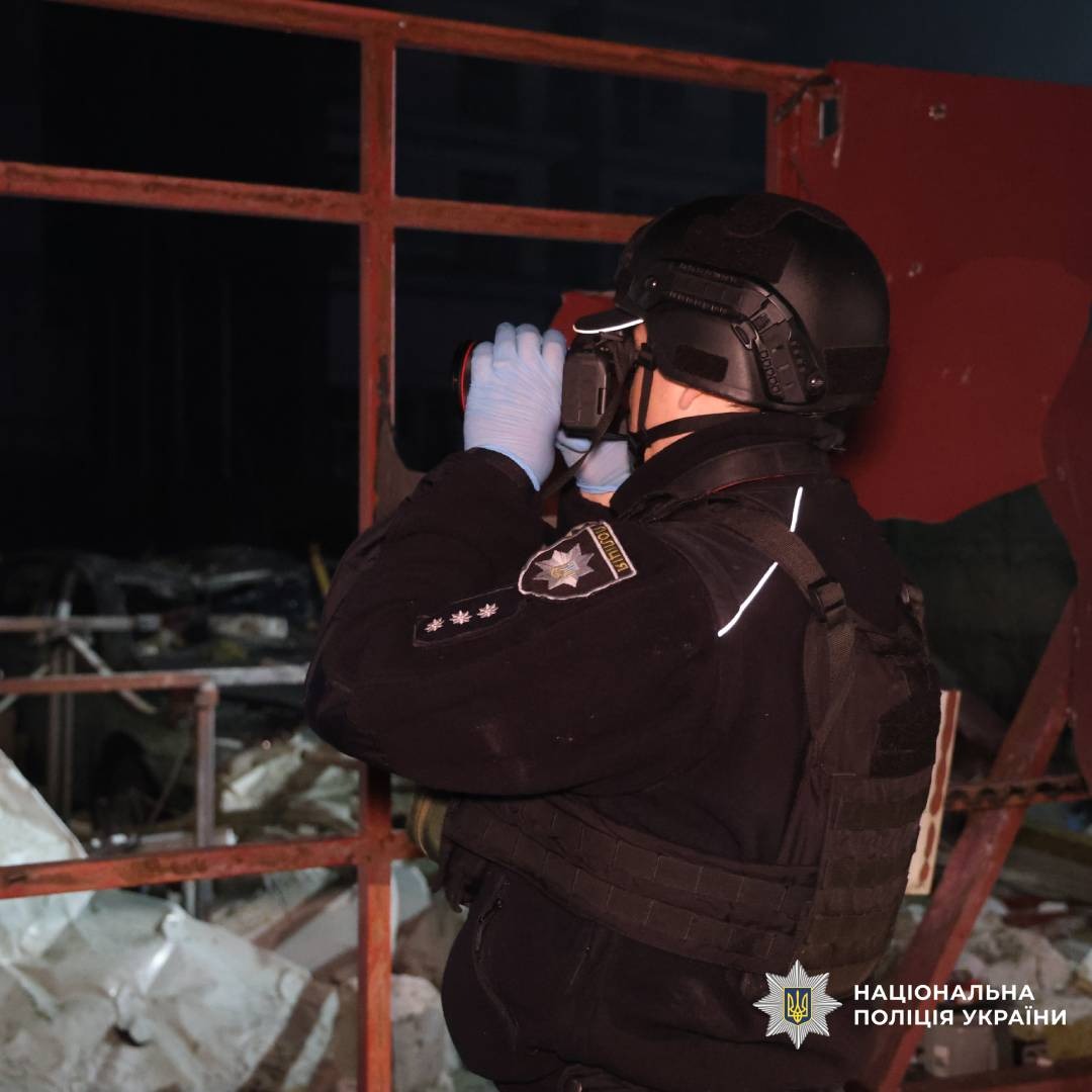 Поліцейський у тактичному спорядженні проводить огляд небезпечної зони за допомогою ручного пристрою. На фоні уламки та пошкоджені конструкції.