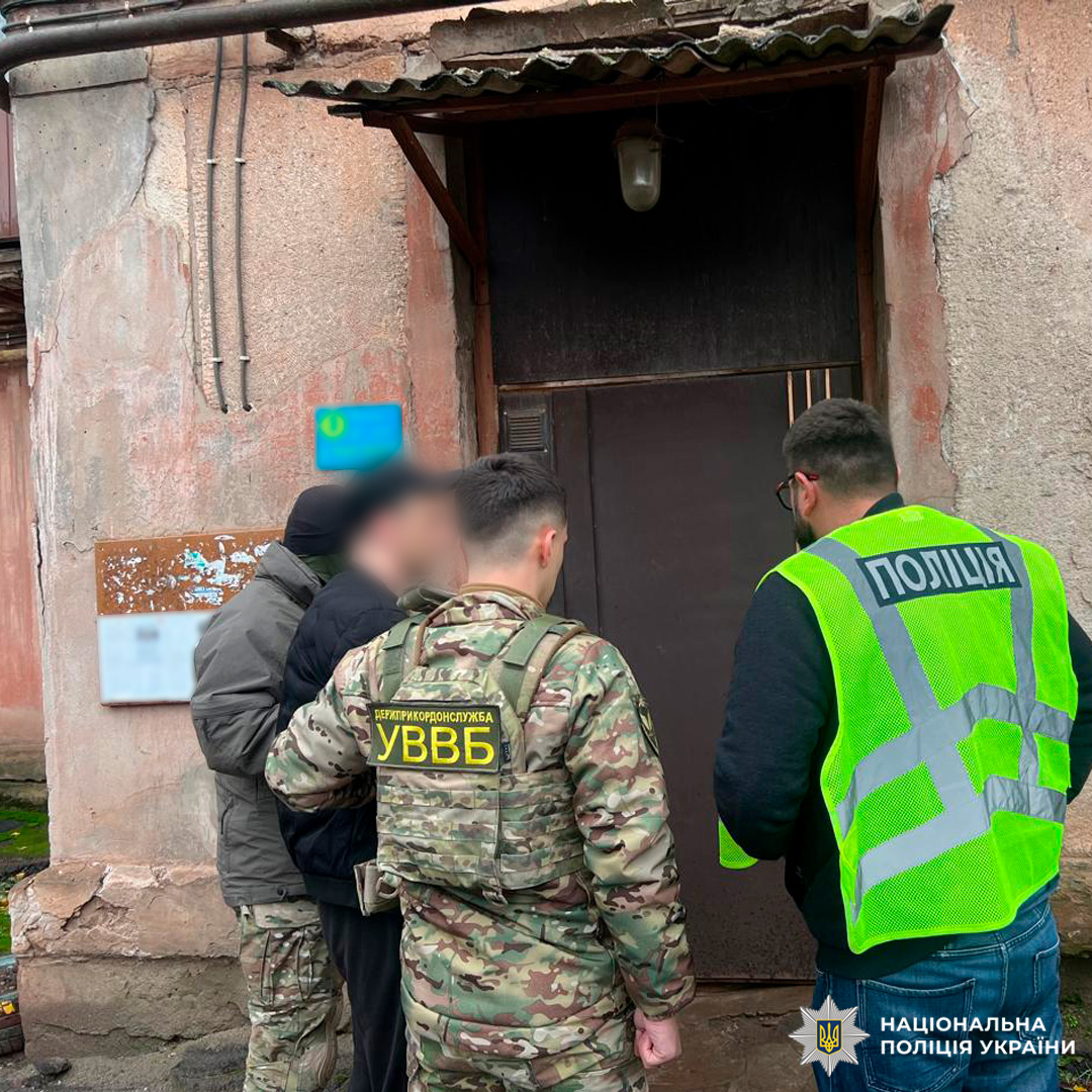 Співробітники Національної поліції України здійснюють оперативні заходи біля входу до будівлі. Один правоохоронець у камуфляжній формі, інший у яскравому жилеті з написом “ПОЛІЦІЯ”. Поруч особа в цивільному одязі.