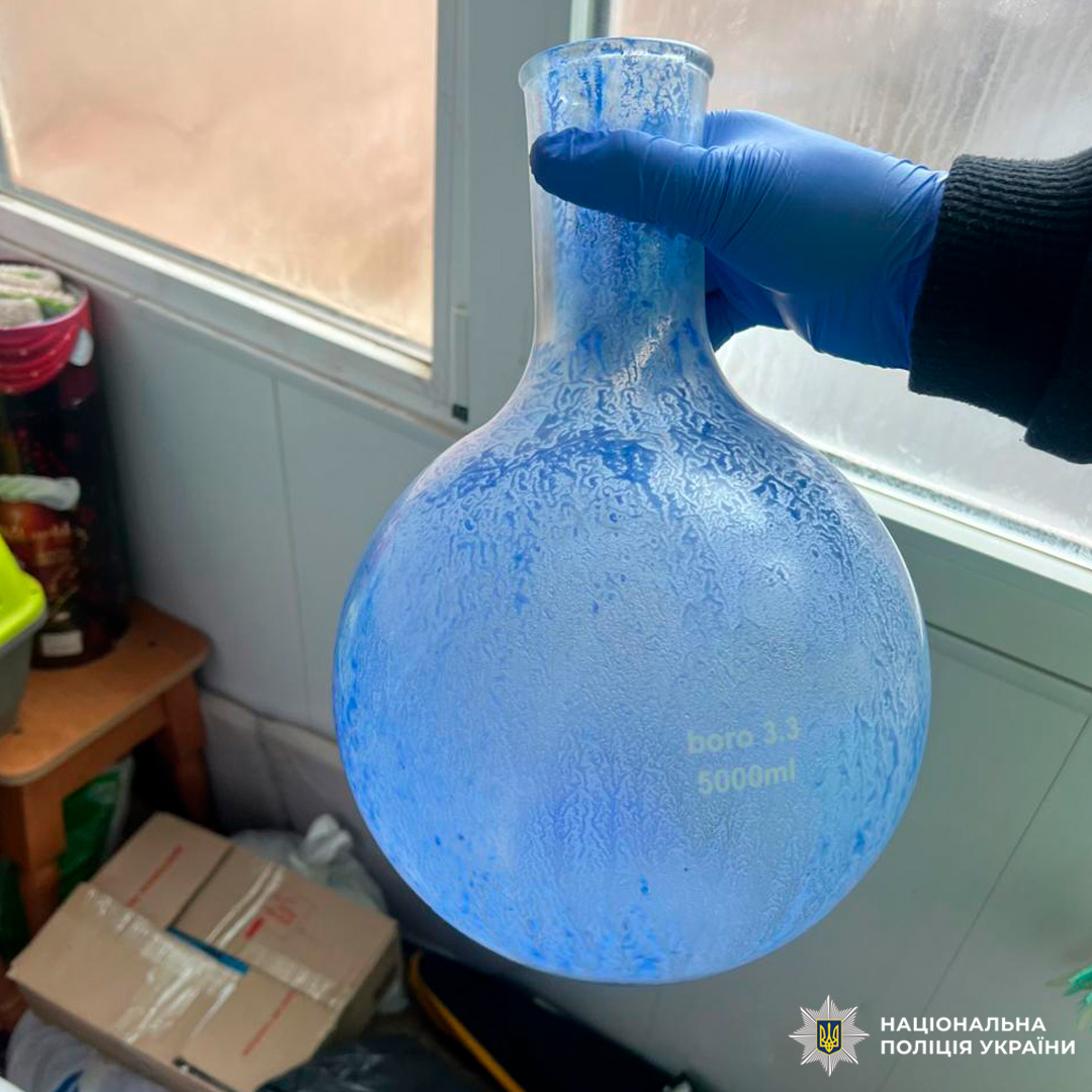 Під час слідчих дій вилучено лабораторне обладнання: особа в захисній рукавичці тримає колбу об’ємом 5000 мл з кристалізованим вмістом синього кольору.