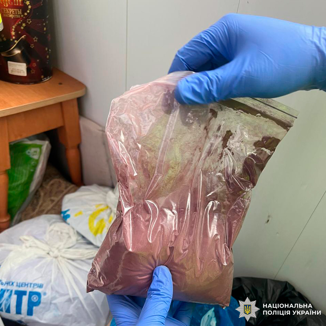 Під час слідчих дій вилучено пакет із порошкоподібною речовиною червоно-коричневого кольору. Особа в захисних рукавичках тримає пакет у приміщенні, де видно дерев’яний стіл та інші пакети на підлозі.