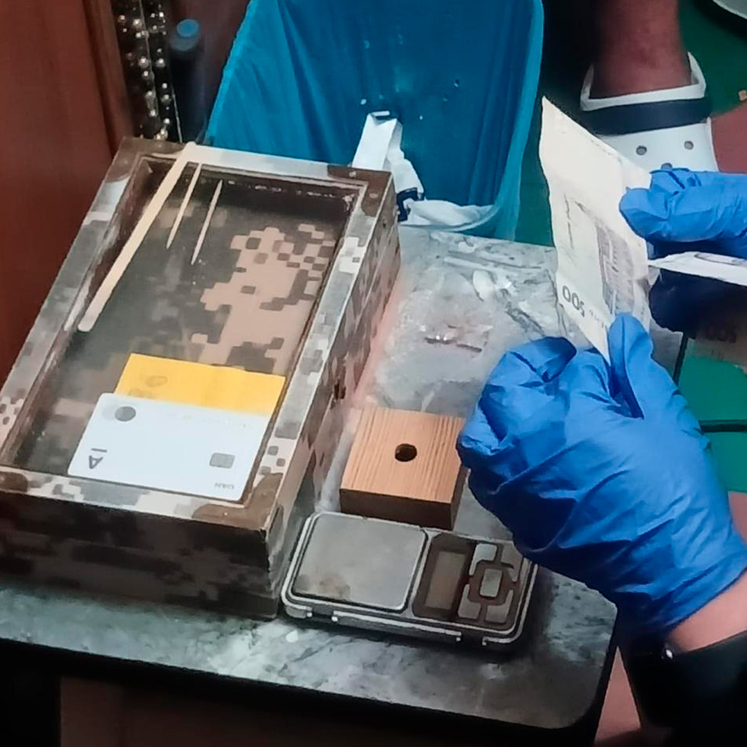 У контрольованих умовах здійснюється обробка речових доказів: особа в захисних рукавичках тримає складений аркуш паперу. На столі камуфльована коробка з предметами, цифрові ваги, дерев’яний блок, пакети та порошкоподібні речовини.