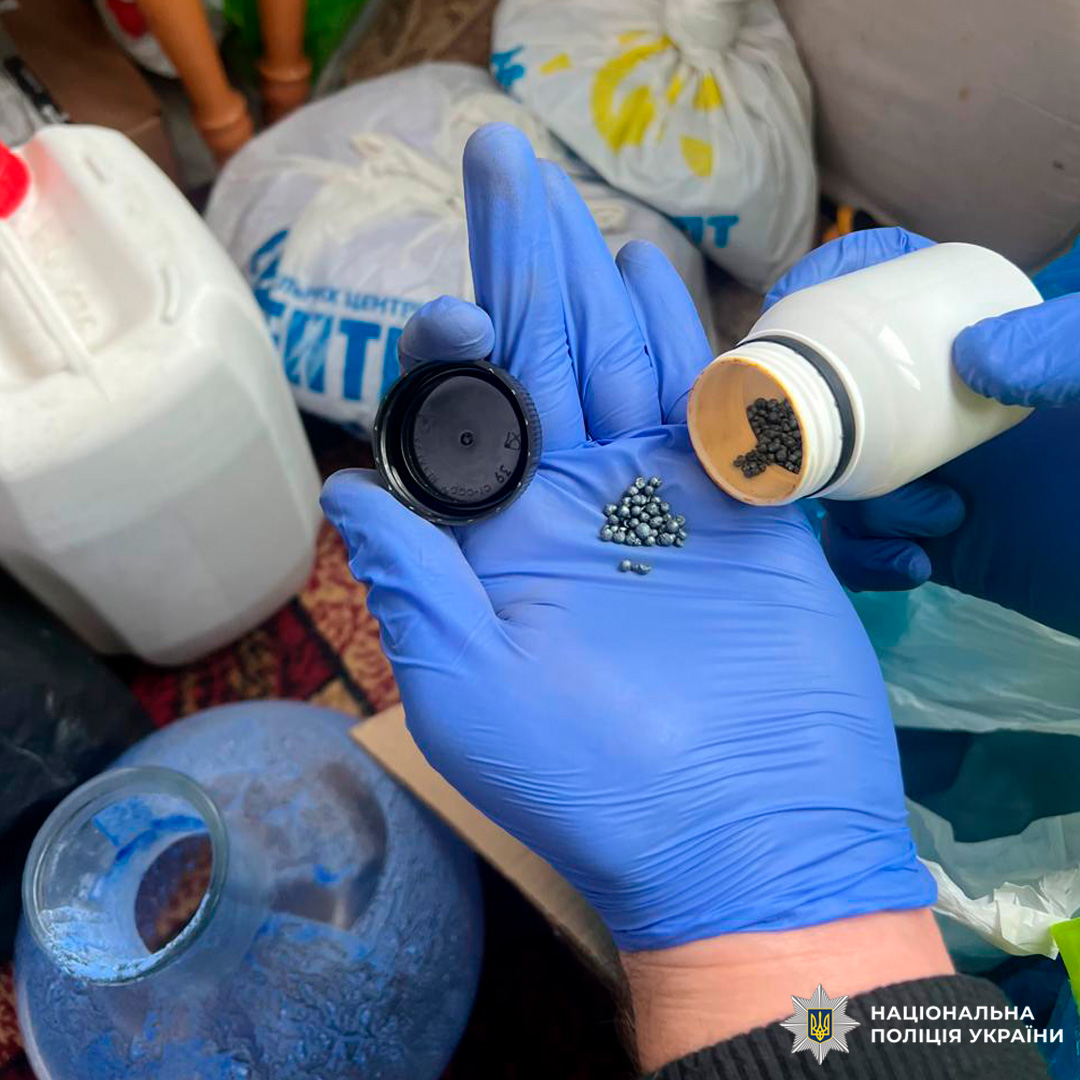 Під час слідчих дій особа в захисних рукавичках висипає металеві гранули з білого контейнера з бежевою кришкою у чорну кришку. На тлі великі ємності, пакети з написами кирилицею та інші предмети.