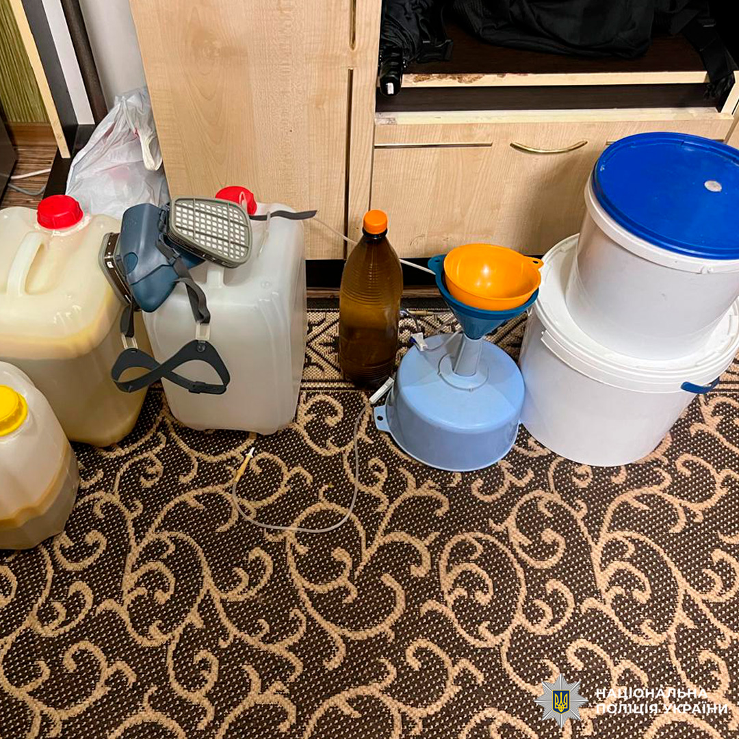 Під час слідчих дій зафіксовано хімічне обладнання та ємності з рідинами, розміщені на килимі в приміщенні. Серед предметів пластикові каністри, пляшка з рідиною, респіратор, відра з кришками, електричний пристрій із лійкою.