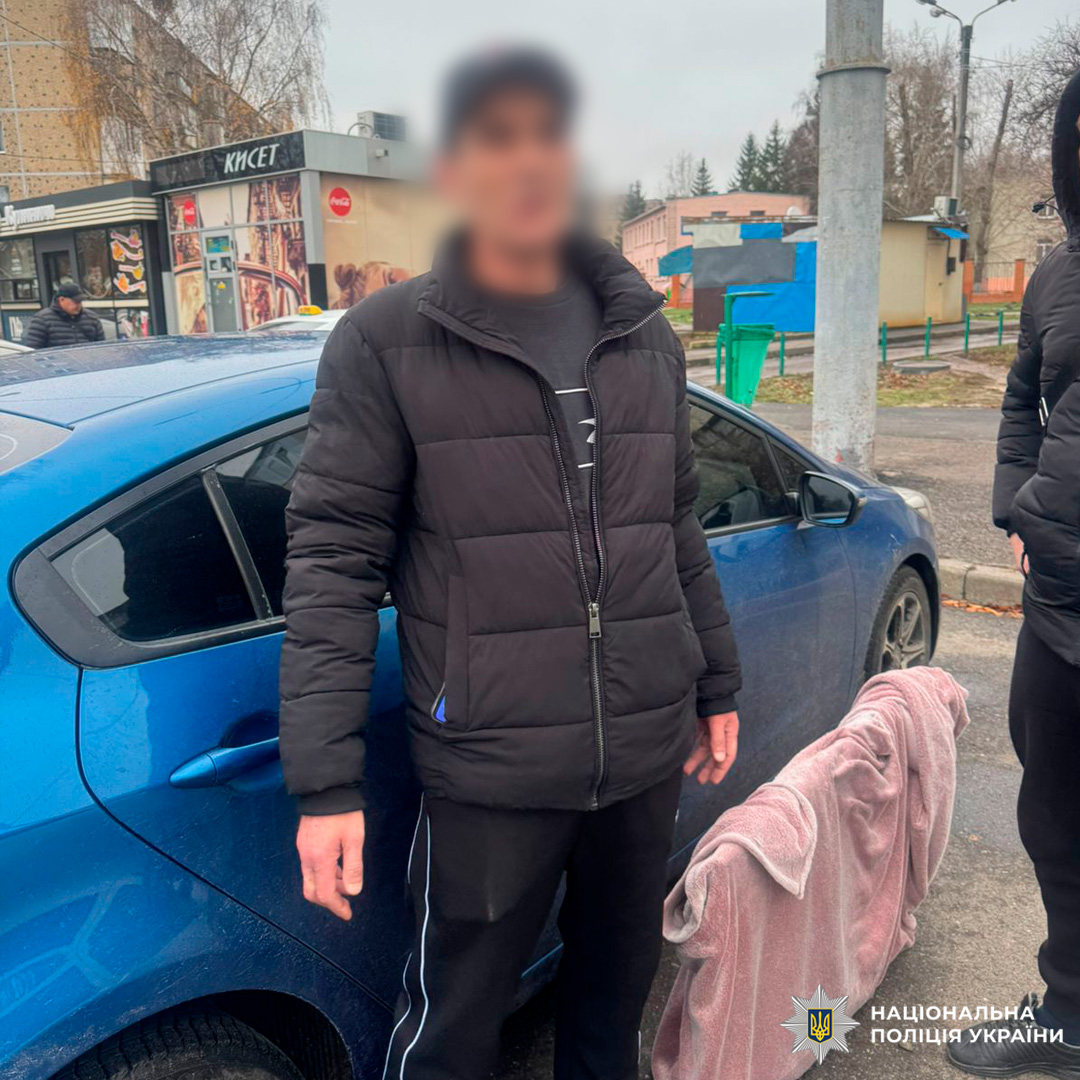 Під час слідчих дій зафіксовано особу в темному одязі біля синього автомобіля в міському середовищі.