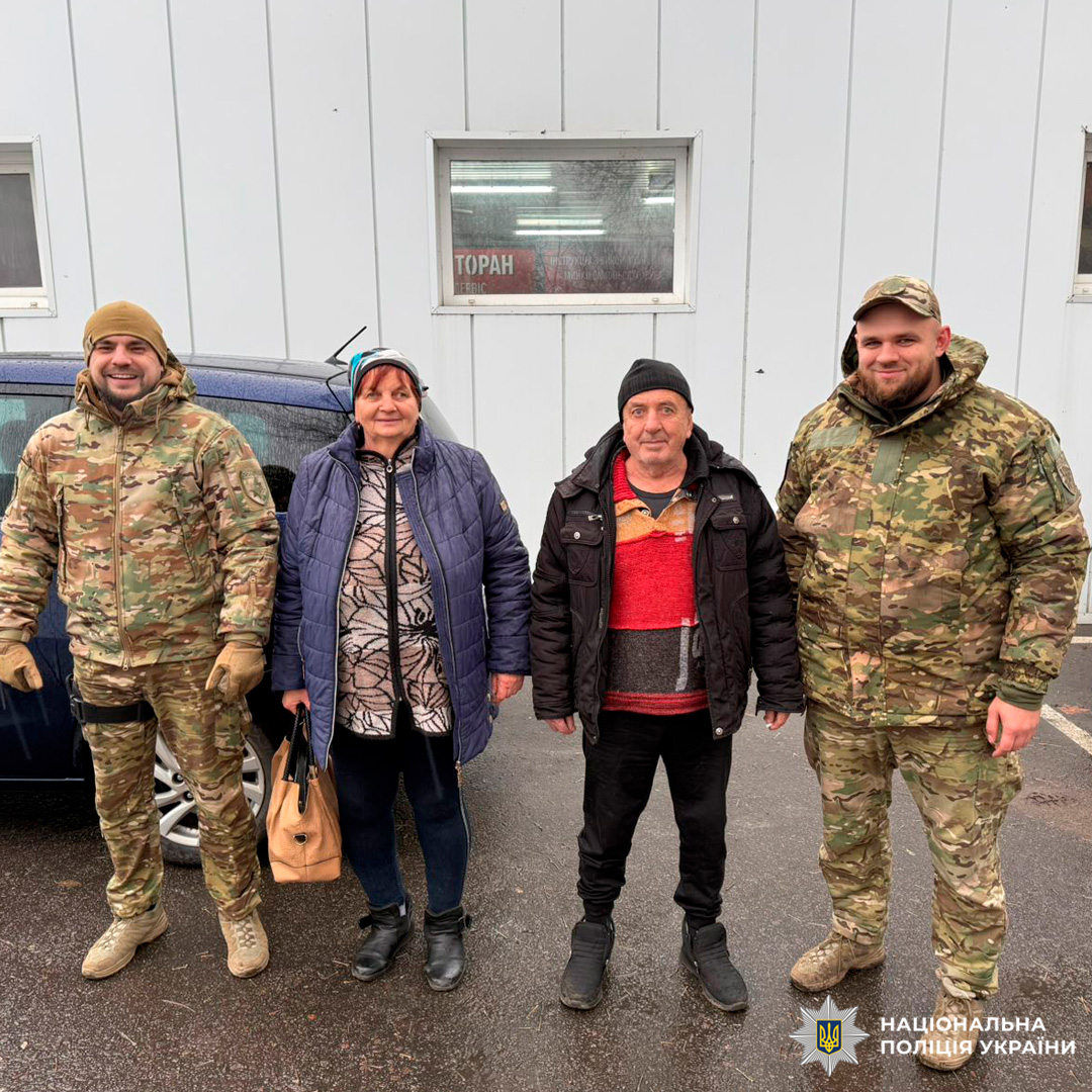 Двоє поліцейских у військовій формі, двоє у цивільному одязі. Жінка в центрі одягнена в синю куртку, сукню з візерунком і чорні легінси, тримає бежеву сумку. Поруч чоловік у чорній куртці та шапці. Поліцейські розташовані по краях групи.