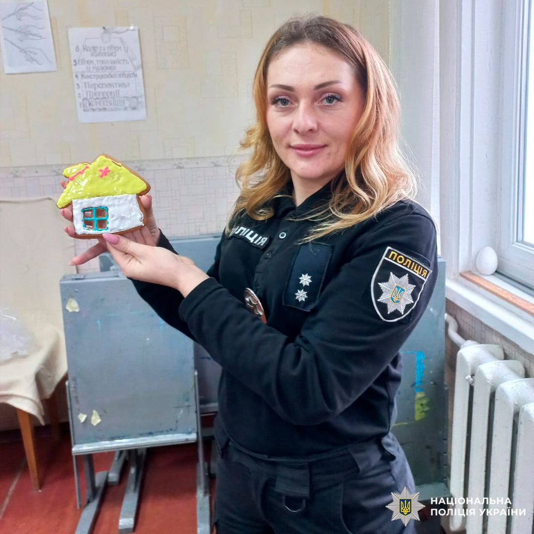 Поліцейська тримає декоративний виріб у формі жовтого будиночка з червоним дахом і синіми вікнами.