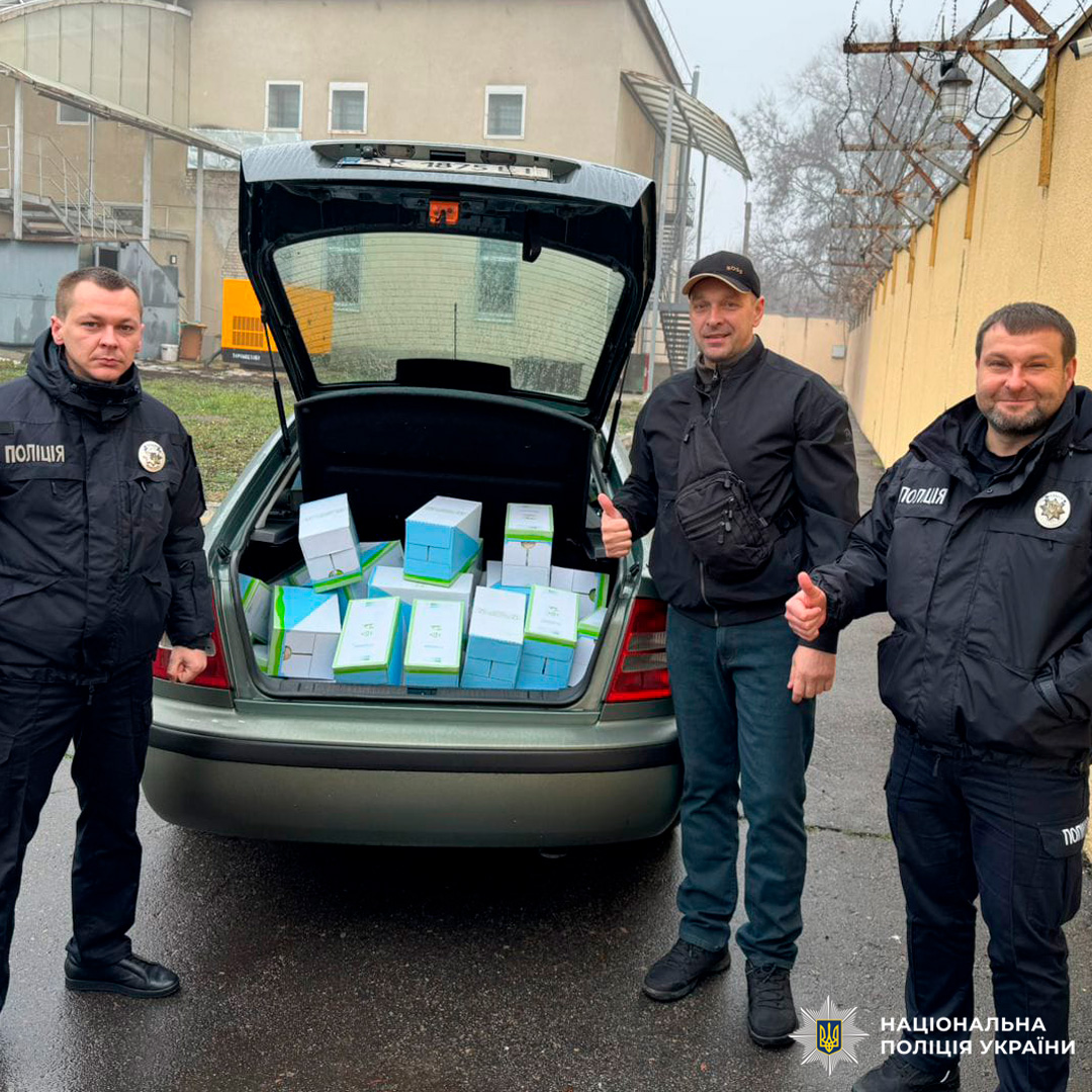 Троє людей біля службового авто з відкритим багажником, заповненим коробками. Двоє у формі поліції, один у цивільному.