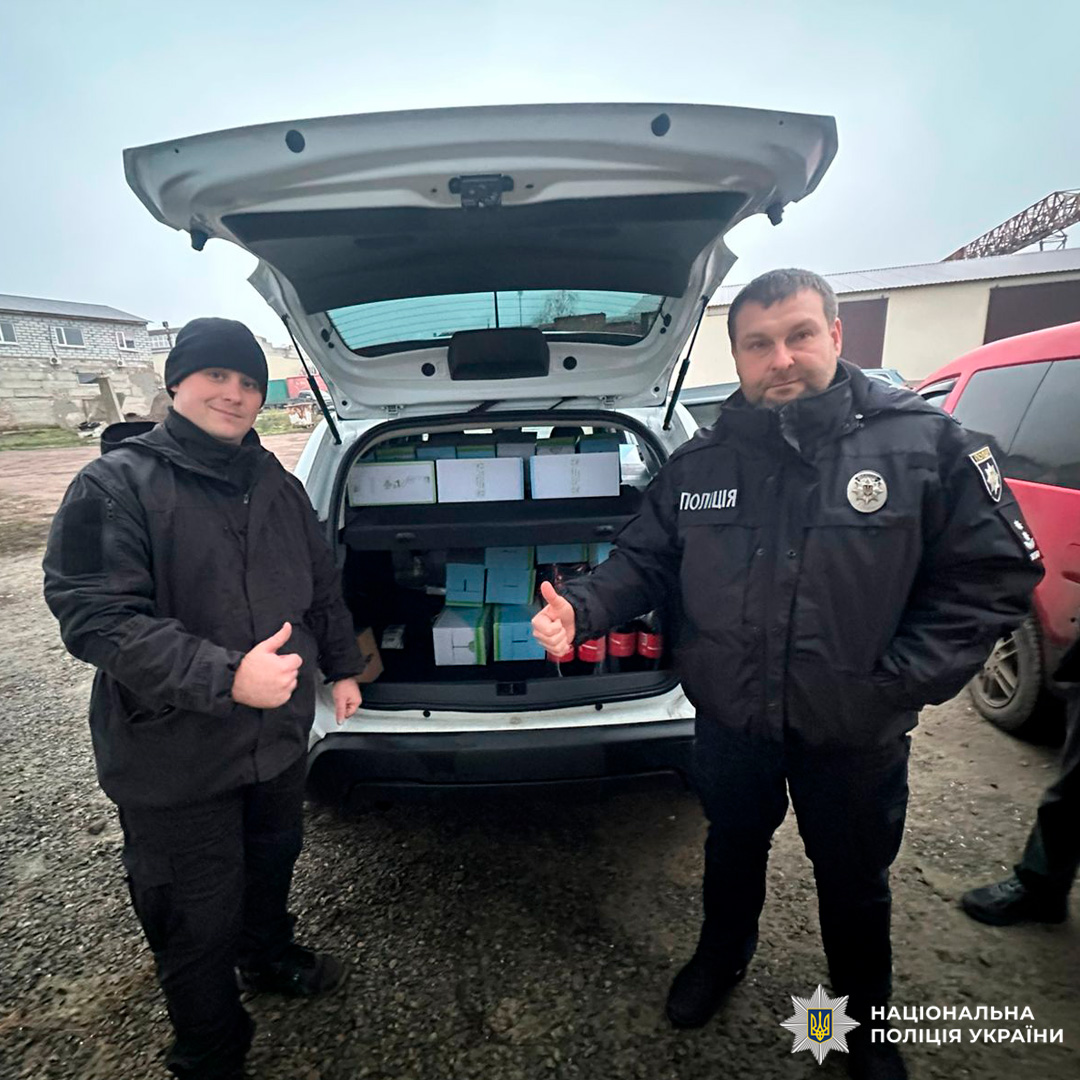 Двоє поліцейських біля відкритого багажника службового авто, заповненого коробками та пляшками. Обидва показують жест «палець вгору».