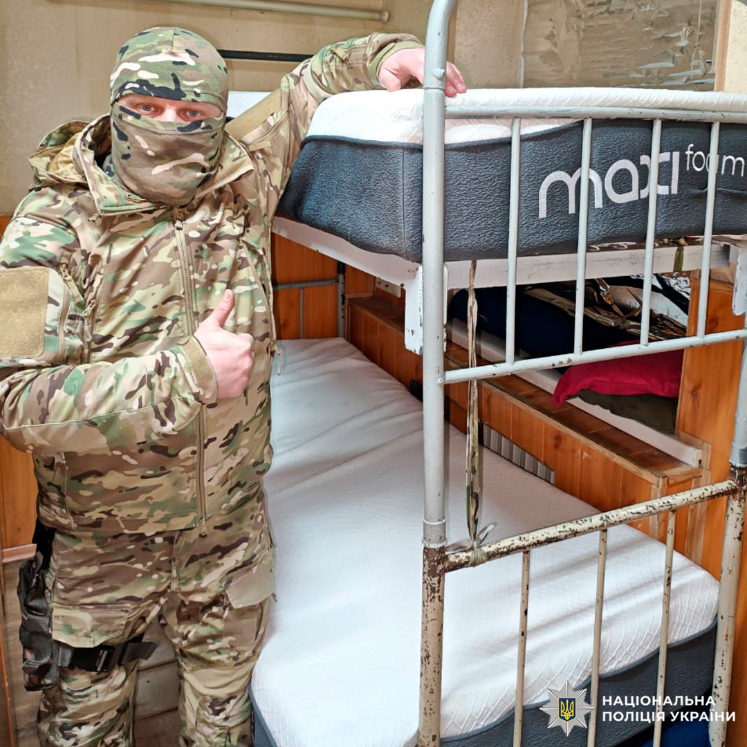 Чоловік у камуфляжній формі біля двоярусного ліжка з охайно застеленими матрацами. Показує жест «палець вгору».