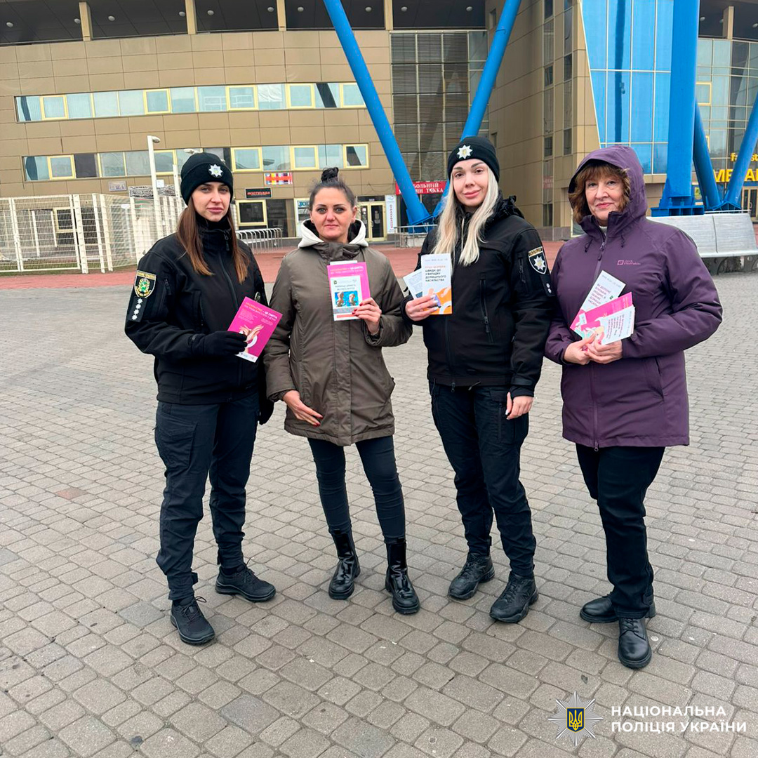 Чотири жінки на вулиці біля сучасної будівлі. Двоє у поліцейській формі, всі тримають інформаційні буклети.