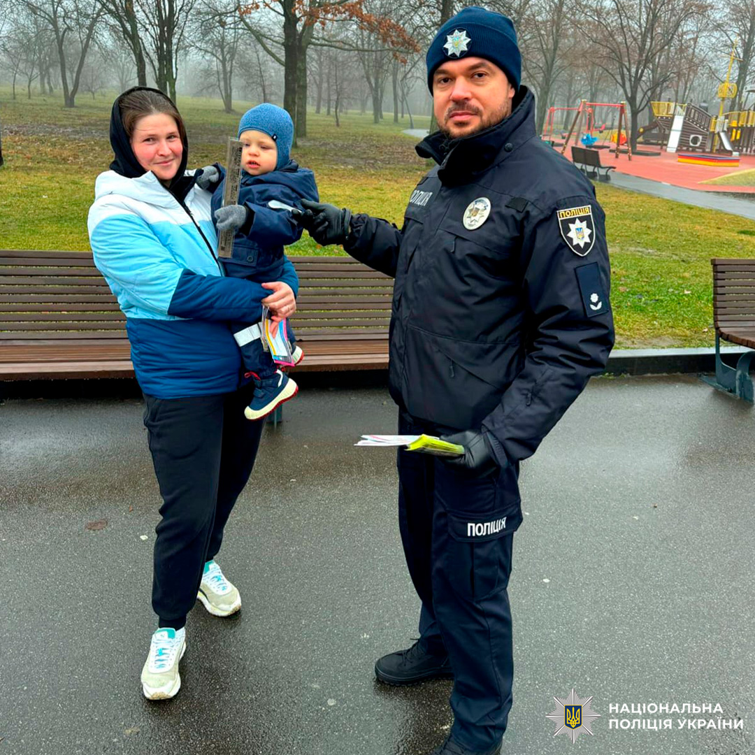 Поліцейський Нацполіції України спілкується з жінкою та дитиною в парку. У руках інформаційні матеріали.