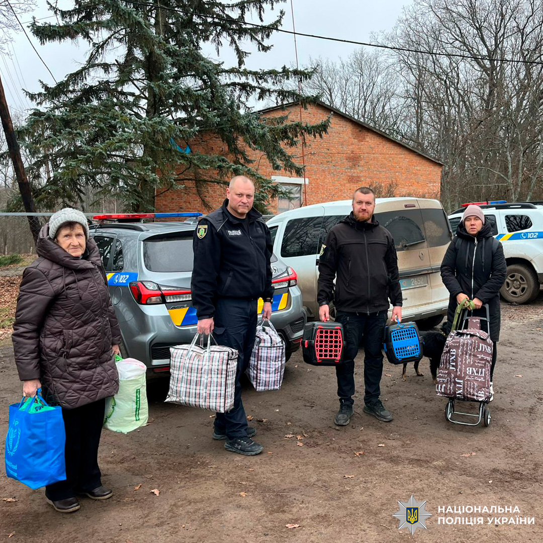 Двоє працівників поліції та двоє цивільних осіб із сумками та переносками для тварин, біля службових автомобілів Національної поліції України. Подія відбувається на тлі цегляної будівлі та дерев без листя, ймовірно, в межах евакуаційного.