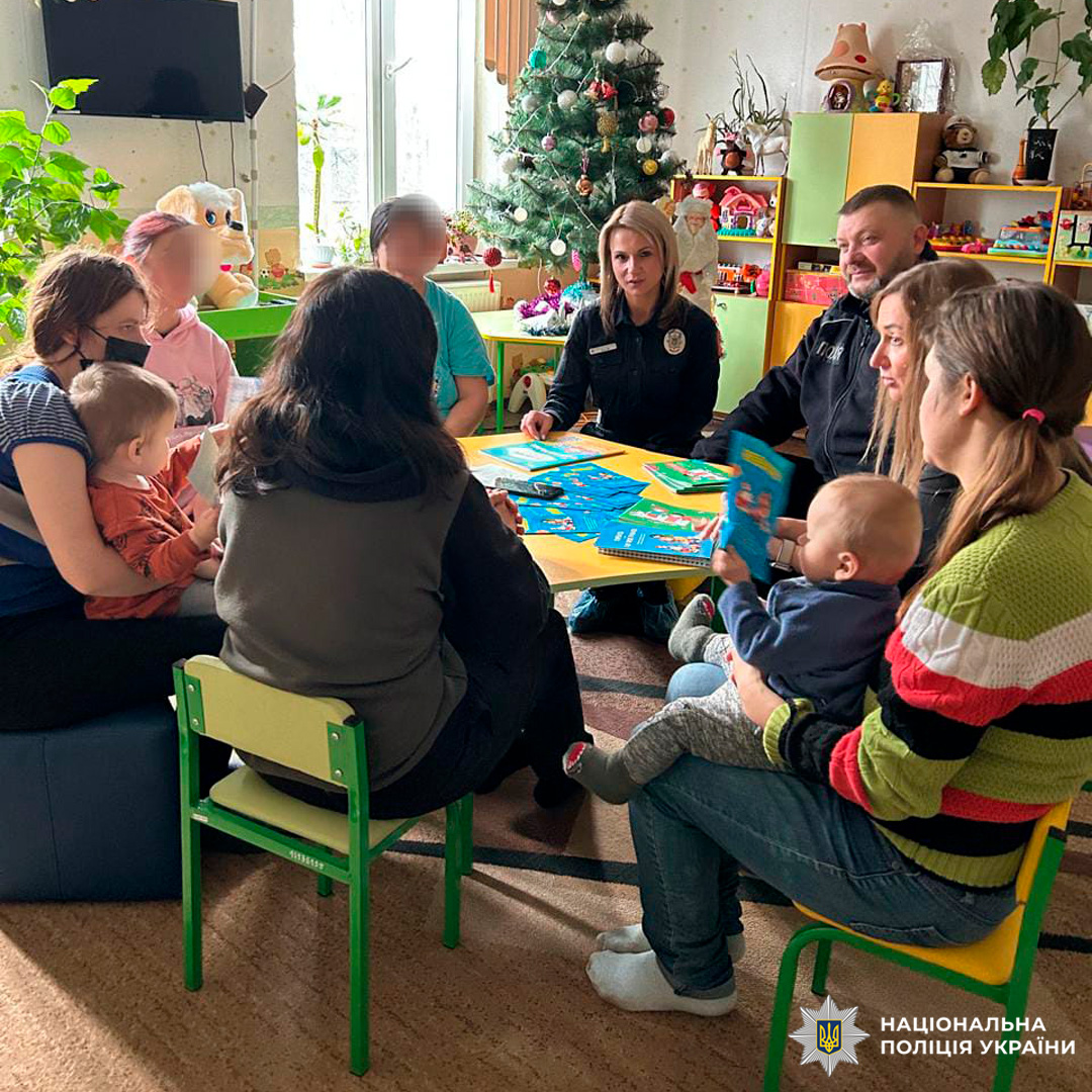 Поліцейські спілкуються з дорослими та дітьми в яскраво оформленій кімнаті. На столі освітні матеріали.