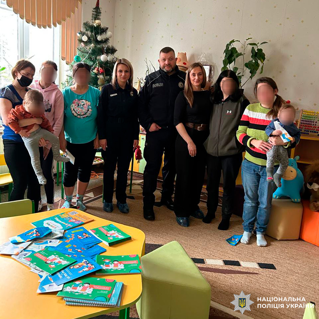 Представники Національної поліції України беруть участь у просвітницькому заході в дитячому закладі. У кімнаті присутні дорослі та діти, інтер’єр прикрашений новорічною ялинкою та іграшками.