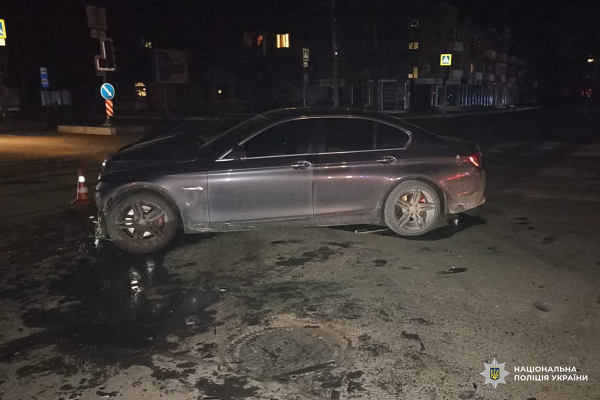 Пошкоджений автомобіль із деформованою передньою частиною стоїть на дорозі вночі; поруч видно дорожній конус та сліди рідини на асфальті.