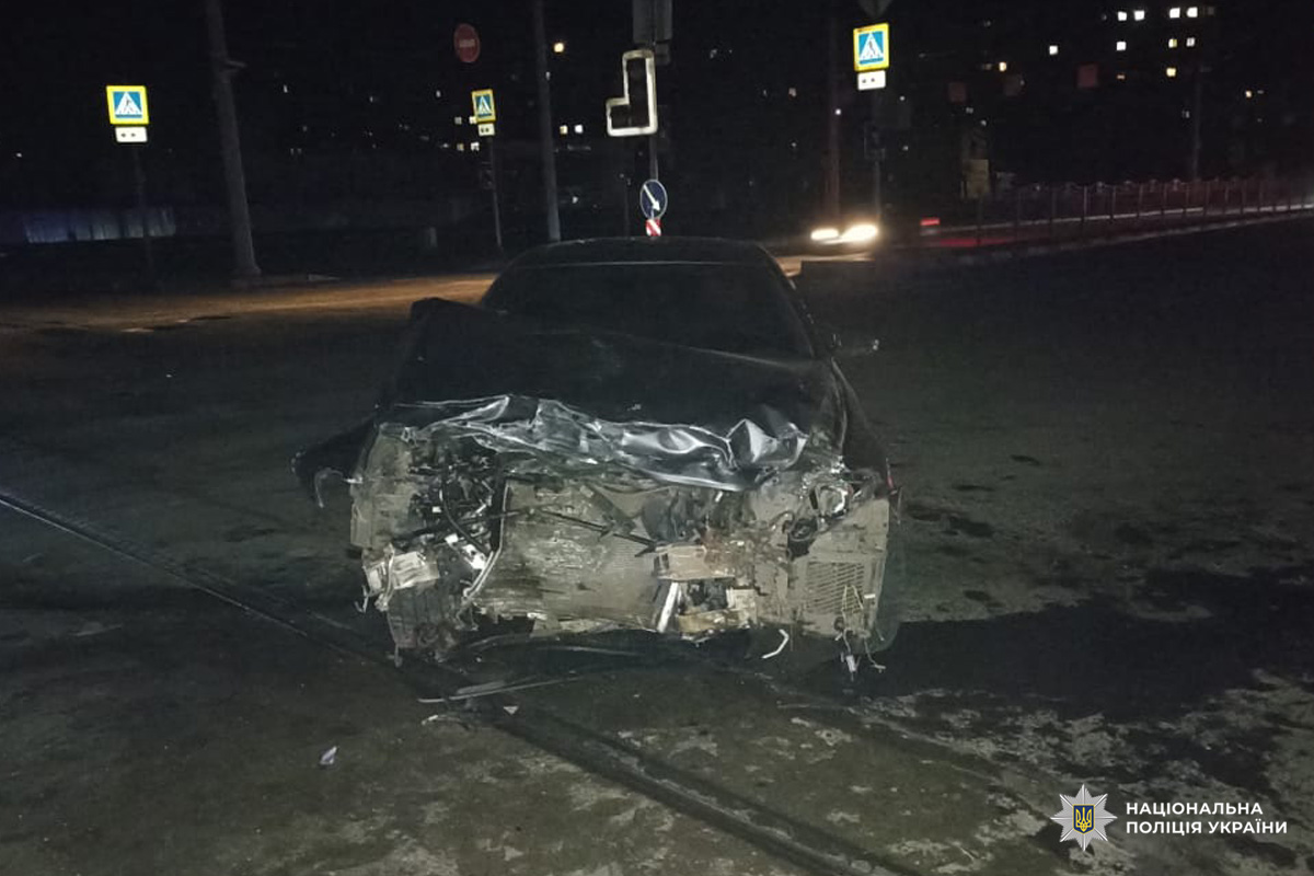 Пошкоджений автомобіль із деформованою передньою частиною стоїть на дорозі вночі; поруч видно дорожній конус та сліди рідини на асфальті.