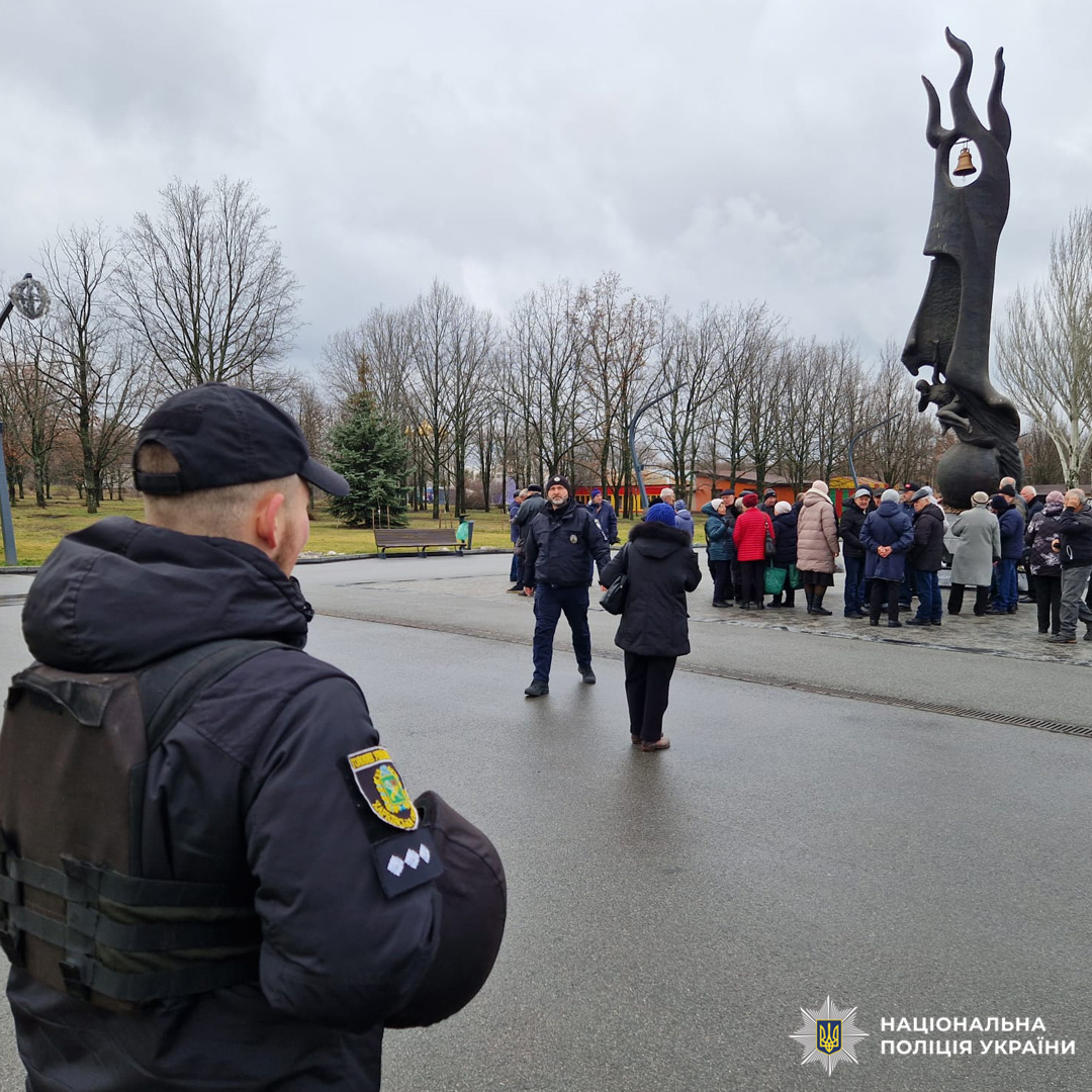 Учасники заходу стоять біля пам’ятника, на передньому плані видно поліцейського в екіпіруванні, навколо дерева без листя.