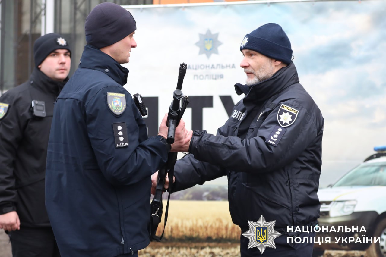 На фото поліцейський офіцер громади, якому керііник вручає помпову рушницю.
