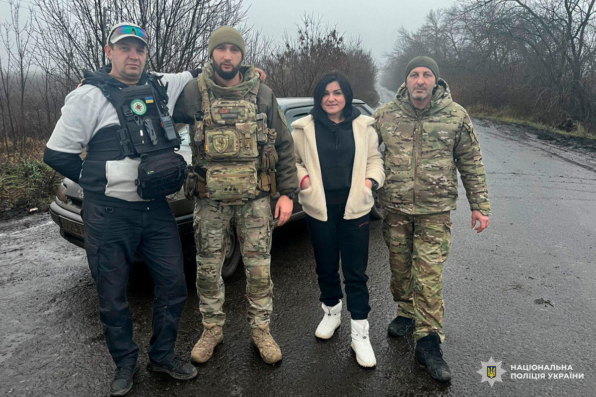 Четверо людей, серед яких правоохоронці, позують для фото на мокрій сільській дорозі біля автомобіля в похмуру погоду.