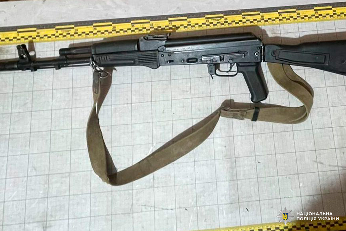 Автоматична гвинтівка з чорним корпусом і коричневим ременем, розміщена на фоні з вимірювальною сіткою та сантиметровою шкалою зверху.