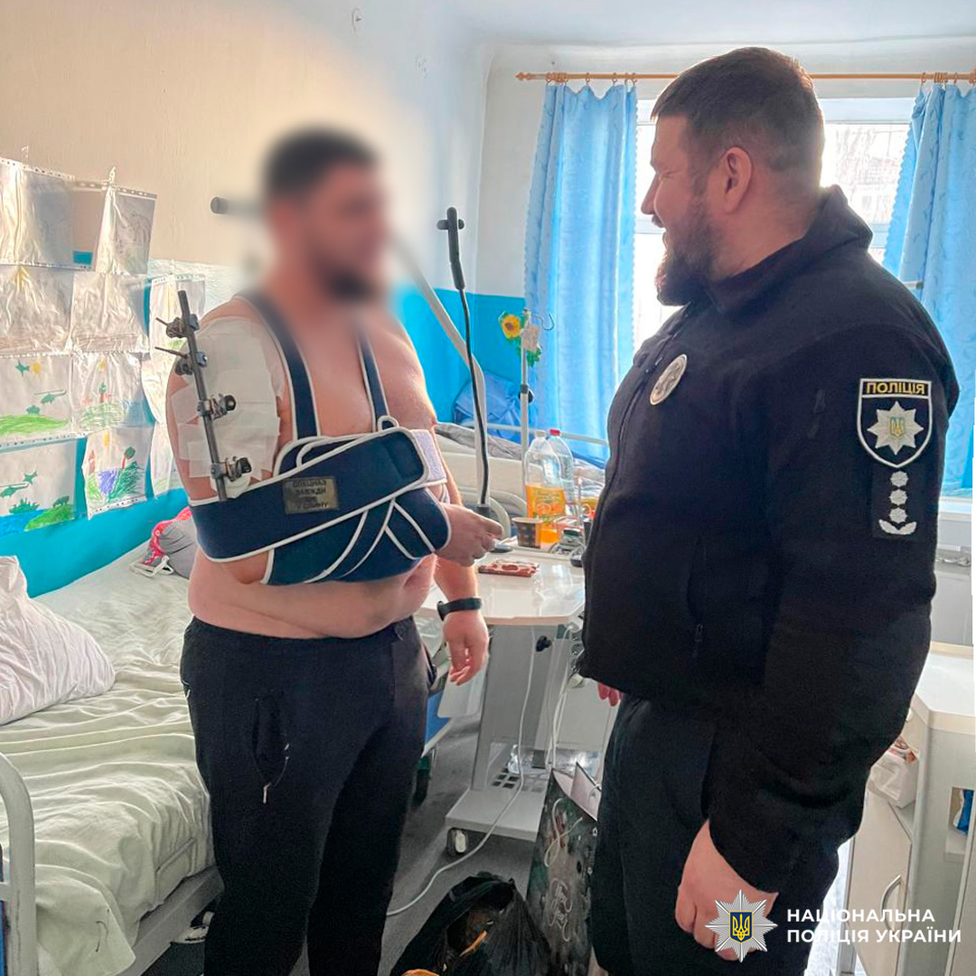 оліцейський підтримує пацієнта з травмованою рукою в лікарняній палаті, серед дитячих малюнків і соняшників.