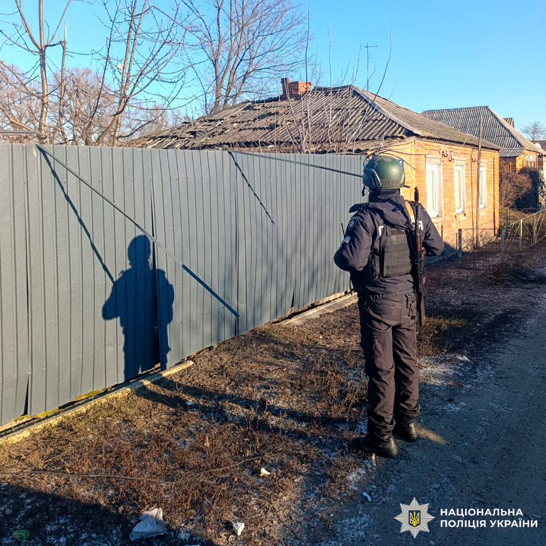 Поліцейський у захисному спорядженні оглядає територію біля житлового будинку з пошкодженим дахом.