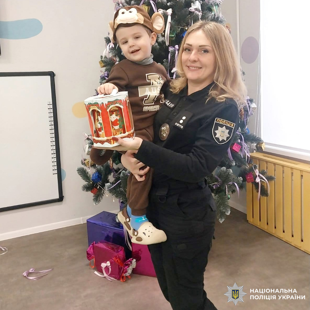 Поліцейська тримає дитину у костюмі мавпочки біля ялинки.
