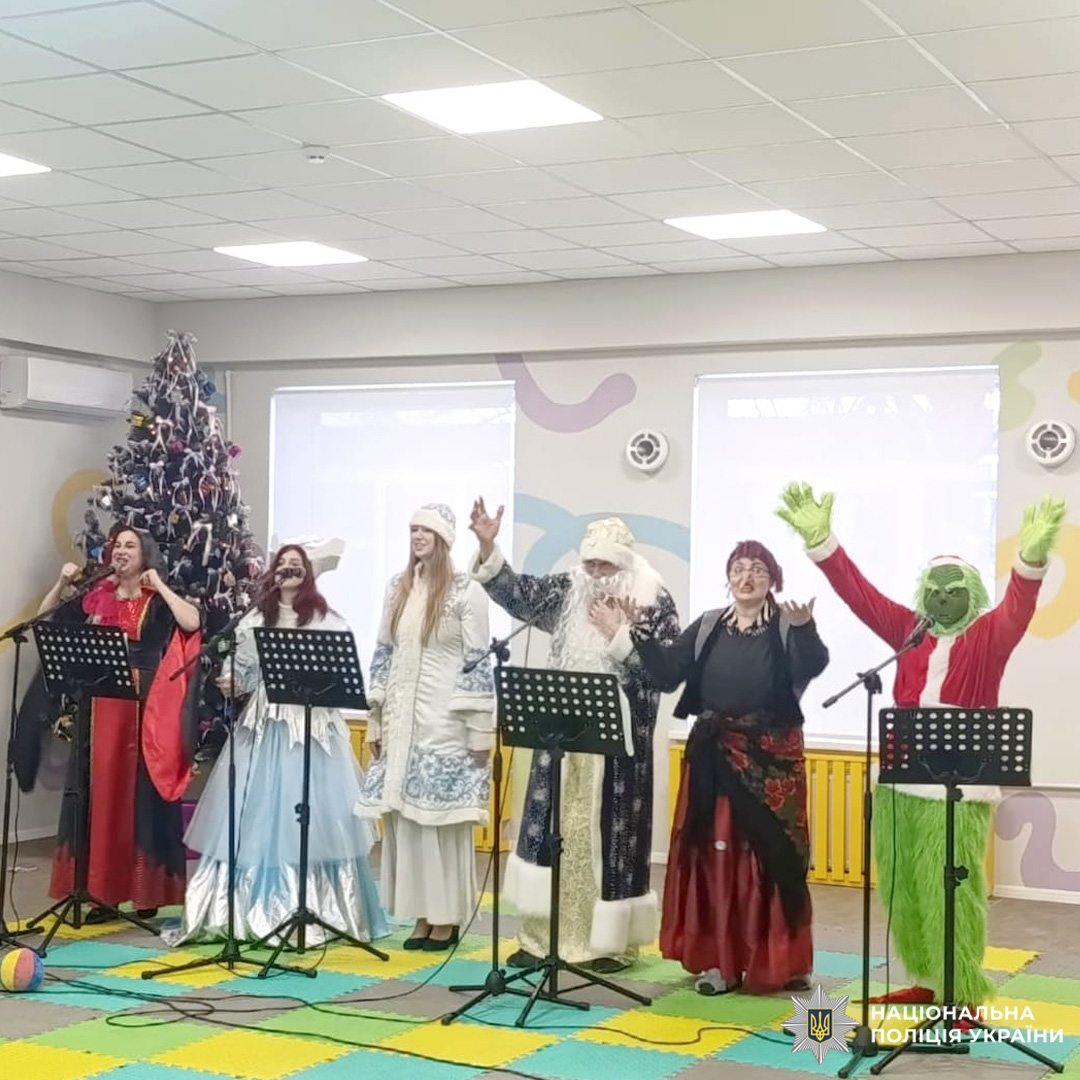 Шестеро людей у святкових костюмах, серед них Дід Мороз та персонаж у костюмі Грінча, виступають перед мікрофонами; на тлі прикрашена ялинка.