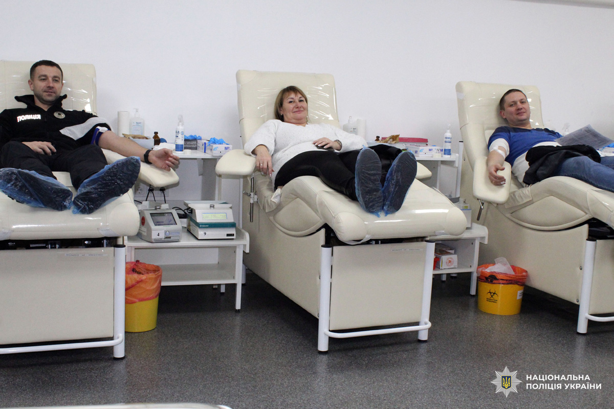 Поліцейський здають кров у клінічному приміщенні; видно апарати для збору крові та медичні матеріали.