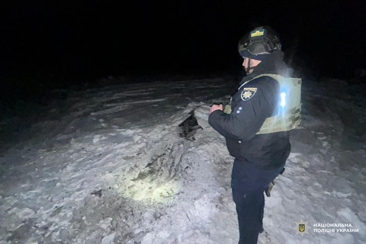 Поліцейський у тактичному спорядженні з каскою та жилетом оглядає вирву в снігу, місце освітлене ліхтарем.