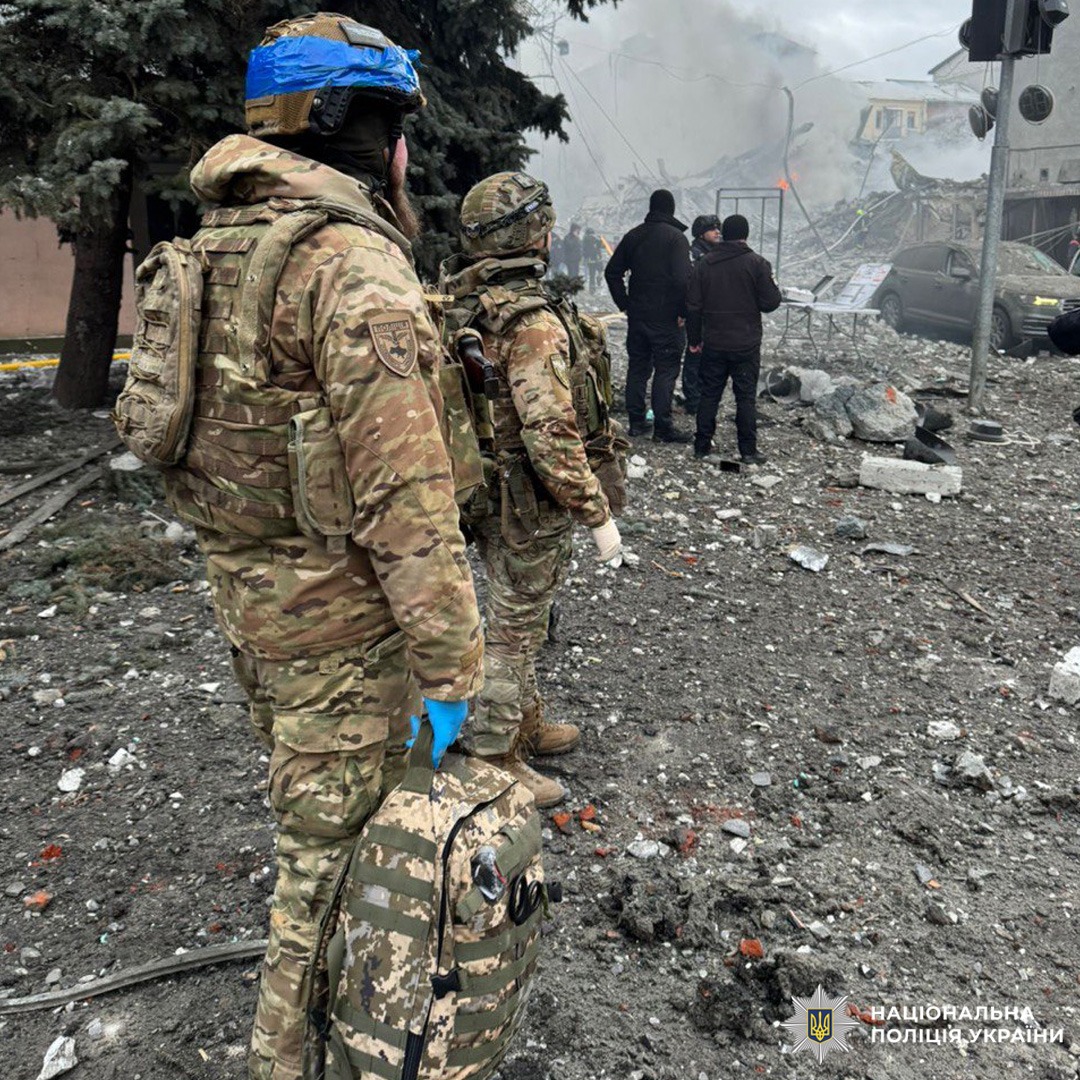 Двоє поліцейських у камуфляжі та спорядженні стоять серед уламків і диму.