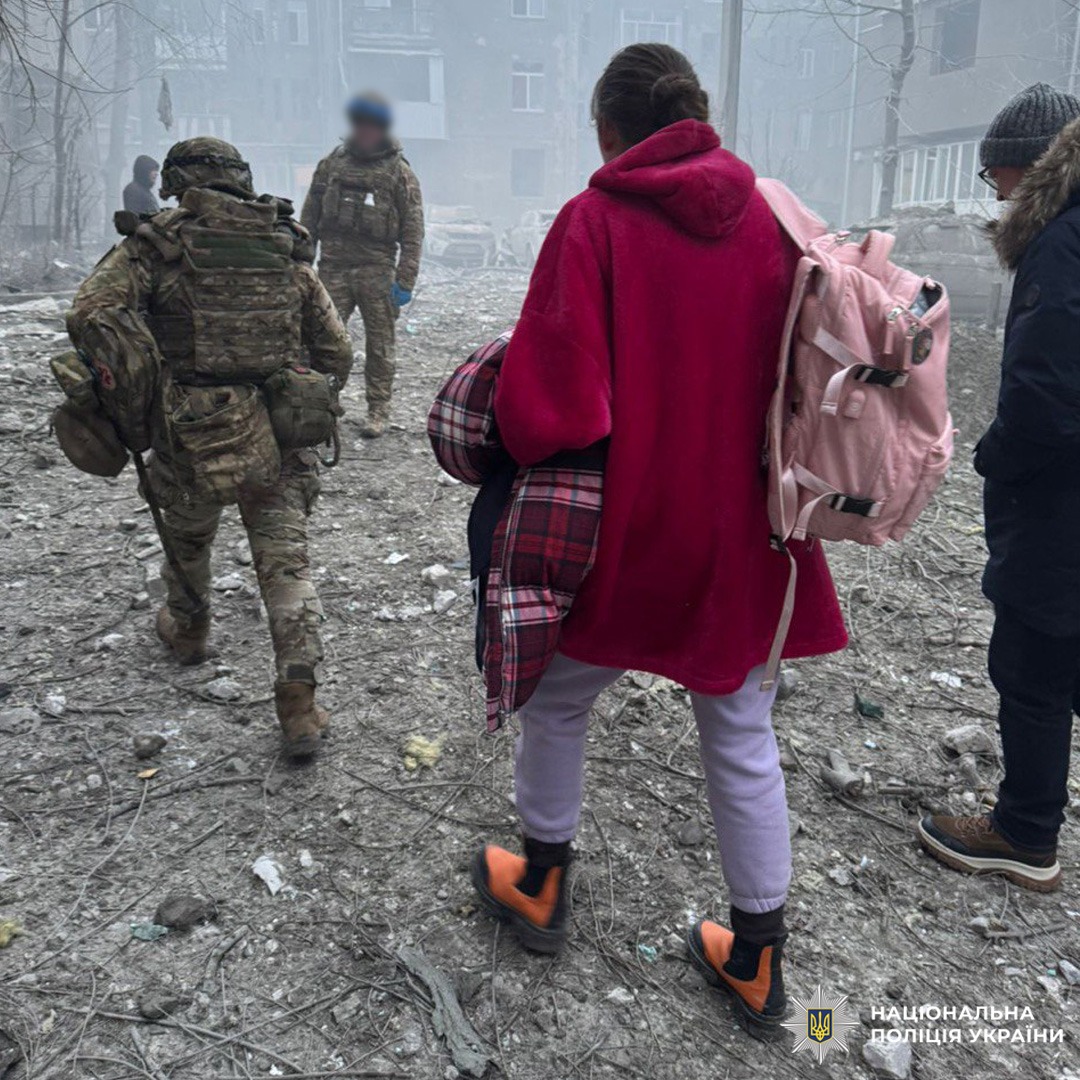 Двоє поліцейських у камуфляжі ведуть групу цивільних через територію з руйнуваннями.