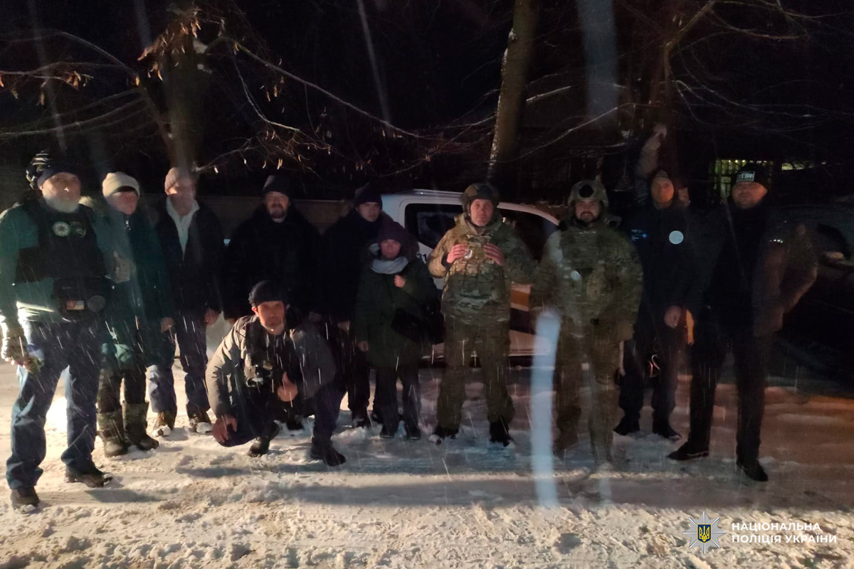 Група людей у зимовому одязі та камуфляжі стоїть біля автомобіля вночі під снігом.