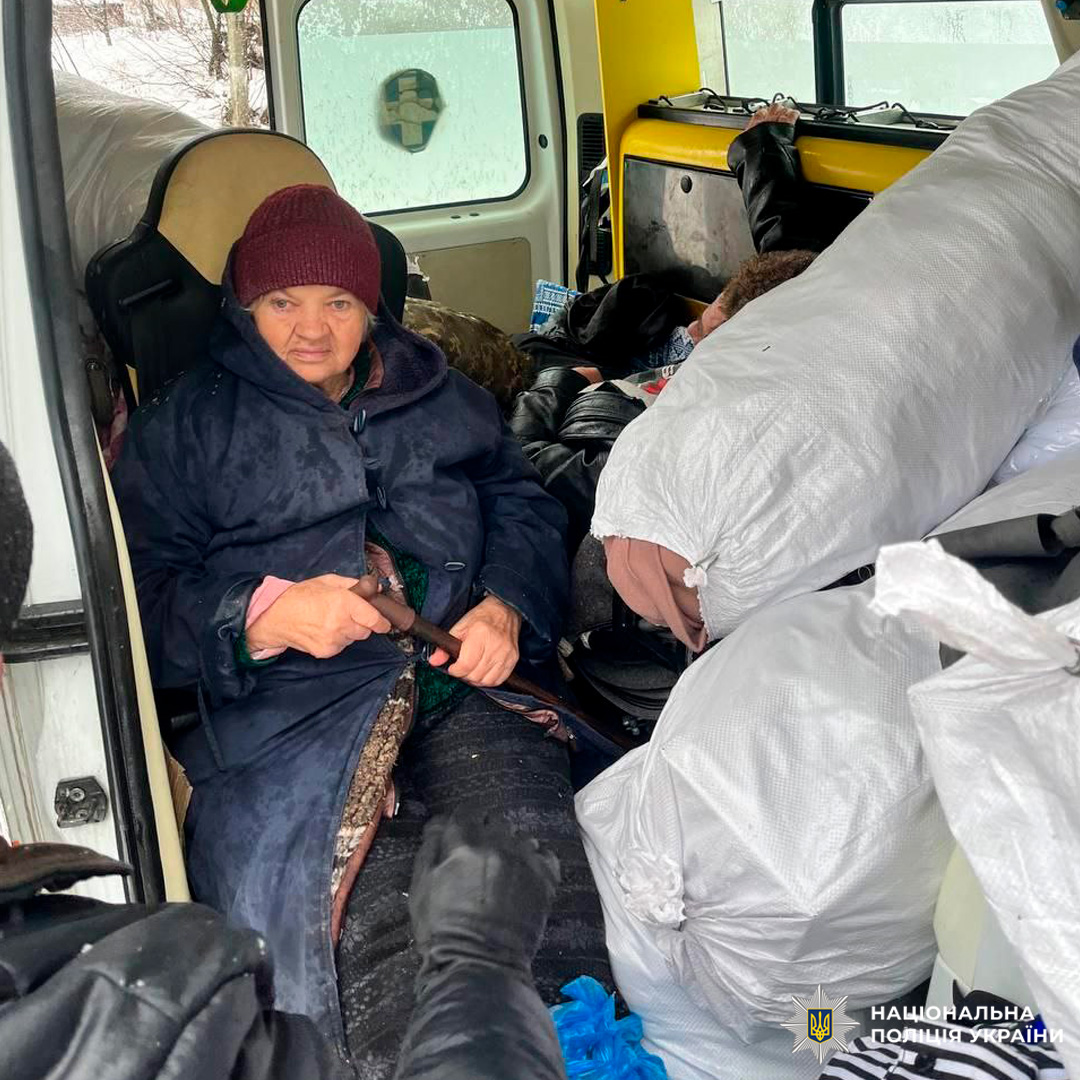 Літня людина у темному плащі та в’язаній шапці сидить із палицею у поліцейському транспорті серед білих мішків.