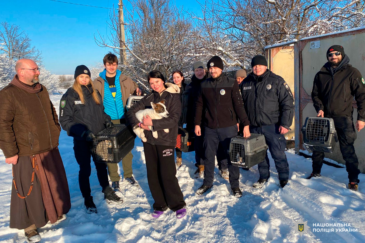 На засніженій місцевості група людей, серед яких є поліцейські у формі, тримає переноски для тварин. Один із учасників тримає маленького собаку. Усі одягнені в зимовий одяг.