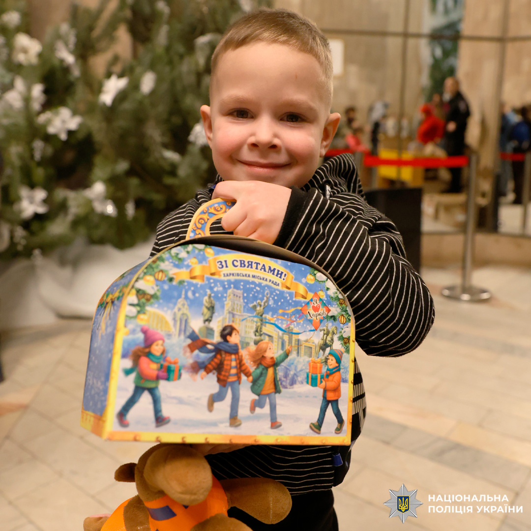 Дитина тримає святковий подарунок та м’яку іграшку біля різдвяної ялинки.