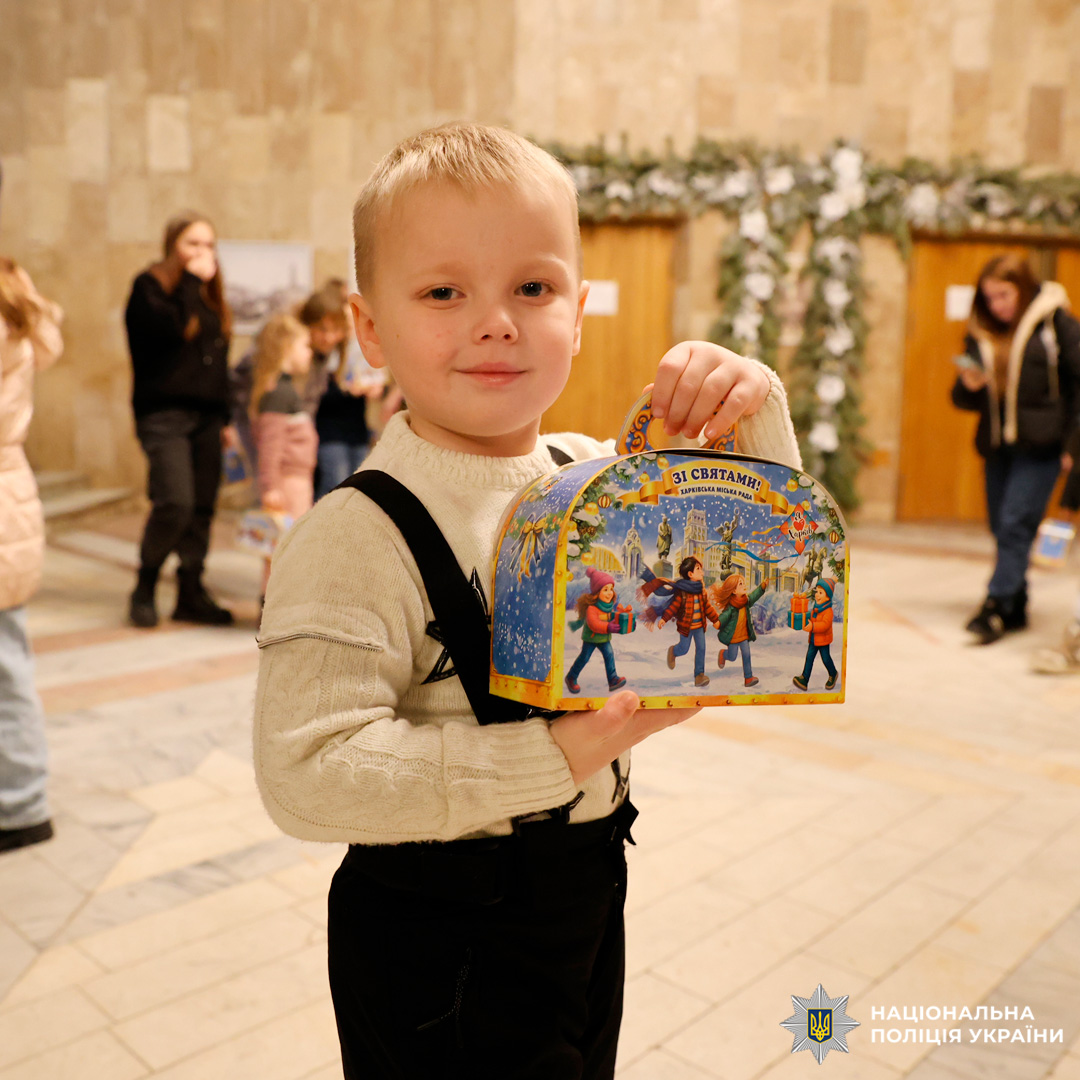 У приміщенні під час святкового заходу дитина тримає яскраву подарункову коробку з написом 'Зі святами!'
